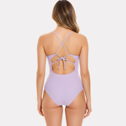 Mujer de espaldas usando traje de baño entero lila con tirantes cruzados y lazo en la espalda.